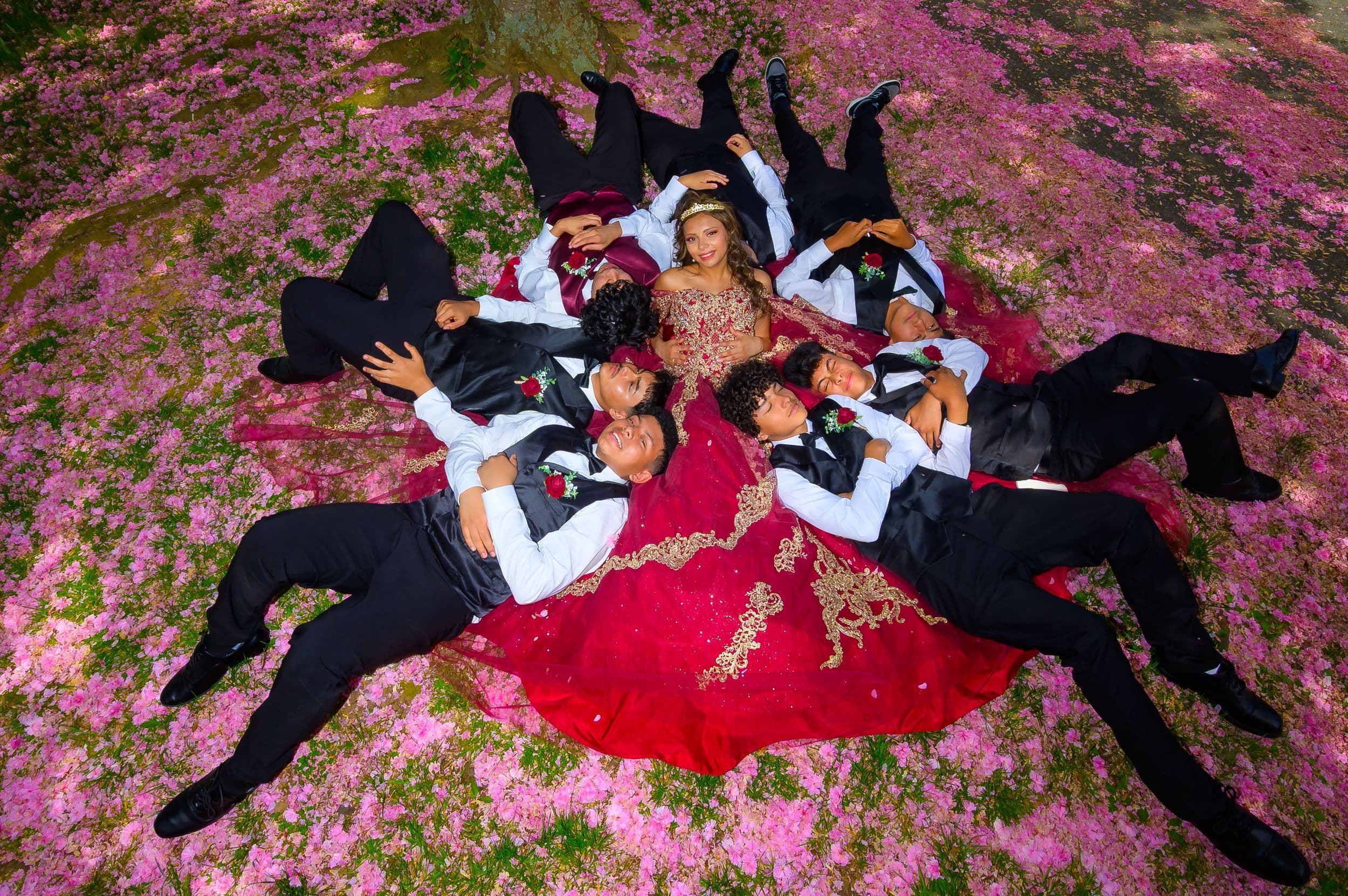 Quinceañera lying in a circle with her chambelanes among pink flower petals